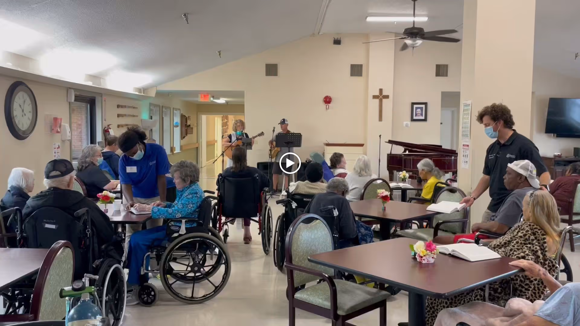 A group of elderly residents, many in wheelchairs, seated around tables in a common area of a senior living facility. Two staff members wearing masks are assisting residents. In the background, two musicians are performing with a guitar and saxophone near a piano. The room has beige walls, a ceiling fan, and a large clock on the wall.