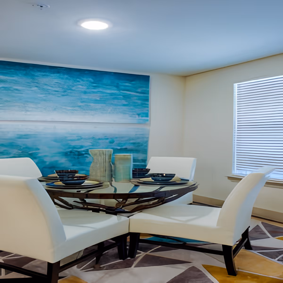 A modern dining area with a round glass table set with plates and bowls, surrounded by four white upholstered chairs. The room features a large blue ocean-themed wall art and a window with blinds, with a patterned rug underneath the table.