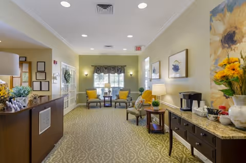 A well-lit, cozy interior common area with patterned carpet, a wooden reception desk on the left, and a granite-topped sideboard on the right holding a coffee maker and decorative items. Two armchairs with yellow cushions and a small table are positioned near a window with curtains at the far end. The walls are painted light yellow and adorned with framed artwork and a large floral painting.