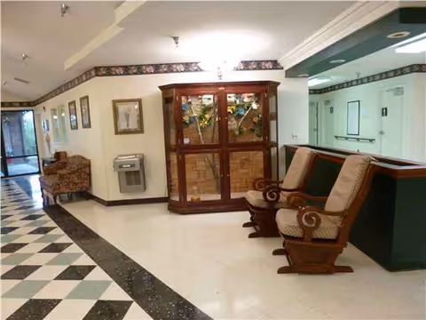 Spacious senior living lobby with checkerboard floor, cushioned wooden chairs, and a glass-front cabinet.