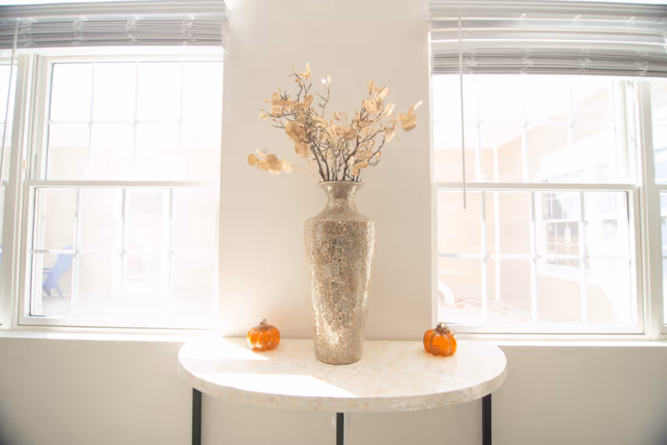 A decorative table with a large ornate vase holding dried branches placed between two windows with white blinds. Two small orange pumpkin decorations are on either side of the vase on the table.