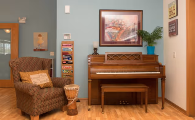 A cozy room with a wooden piano and matching bench against a light blue wall. Above the piano is a framed floral artwork and a potted plant in a blue pot. To the left of the piano is a patterned armchair with a pillow that reads 'Homestead Living Our Friends,' and a small drum on the floor next to it. A wooden door with frosted glass panels is partially visible on the far left, along with a rack holding magazines or brochures.