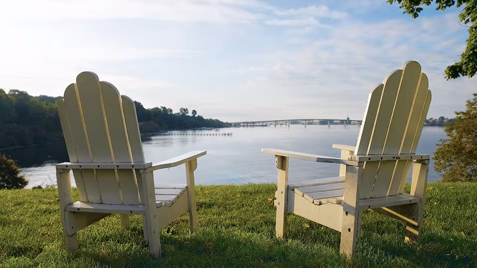Two white wooden Adirondack chairs on a grassy hill overlooking a calm body of water with a bridge in the distance and trees on either side under a partly cloudy sky.