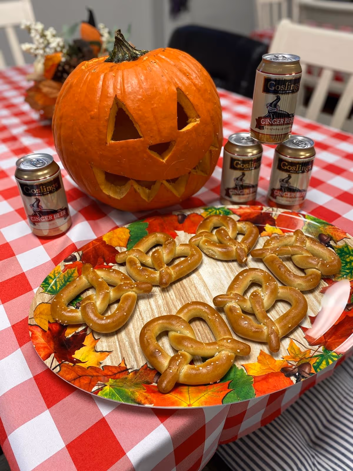A carved jack-o'-lantern and cans of ginger beer on a red-and-white checkered table beside a fall-themed plate of soft pretzels.