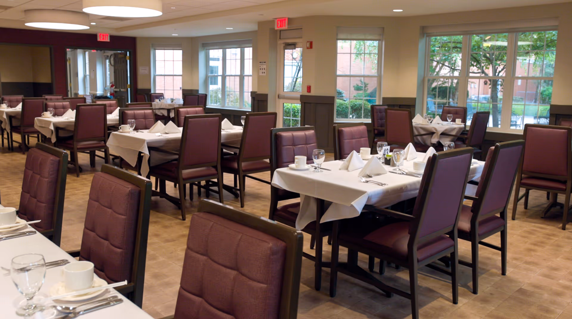 Dining room with neatly set tables, burgundy upholstered chairs, and large windows.