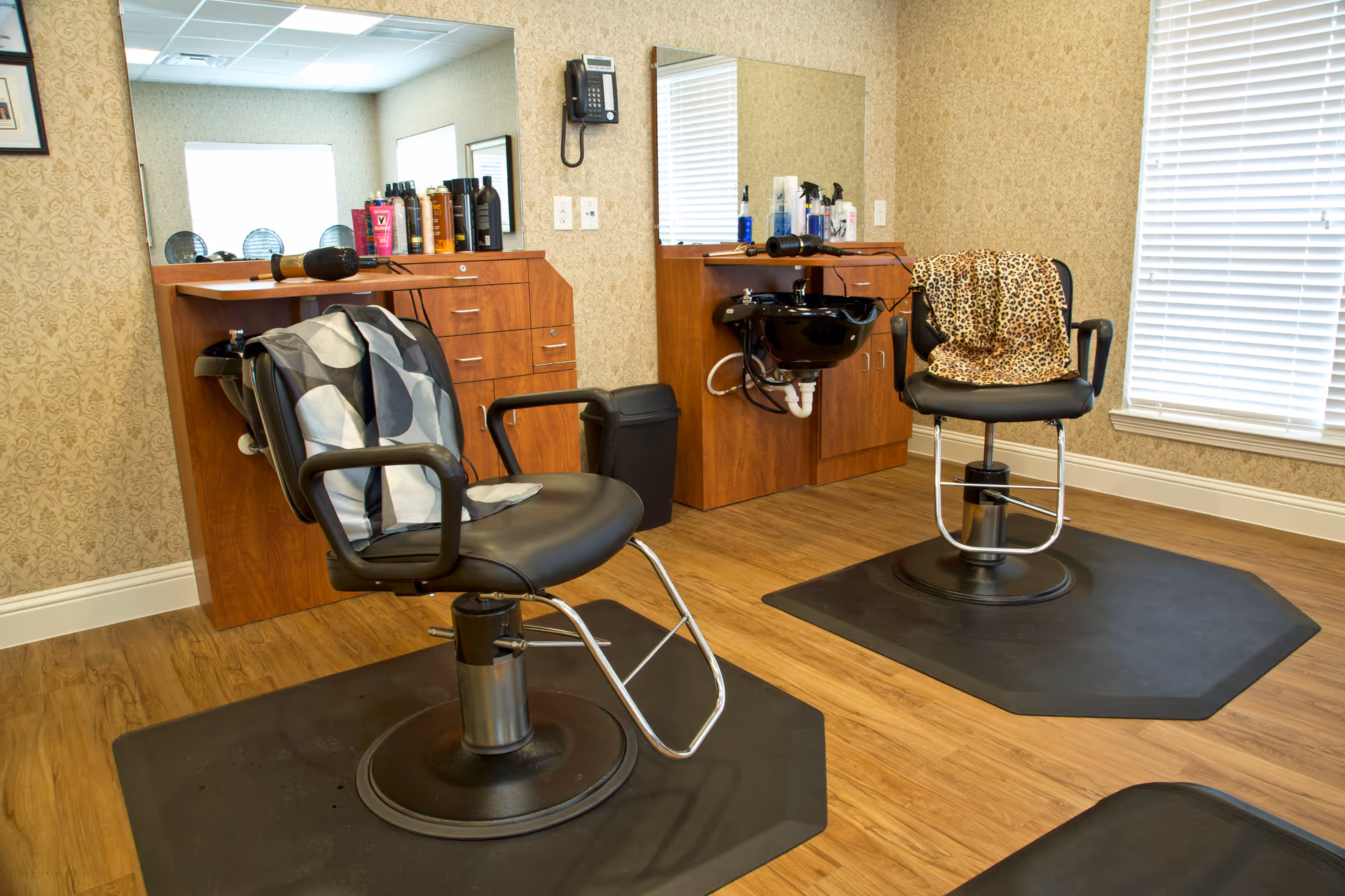 Interior view of a hair salon area with two black salon chairs on black mats, each with a patterned cape draped over the back. Behind the chairs are wooden cabinets with hair care products, a mounted telephone, and a black wash basin. The room has beige patterned wallpaper and a window with white blinds.