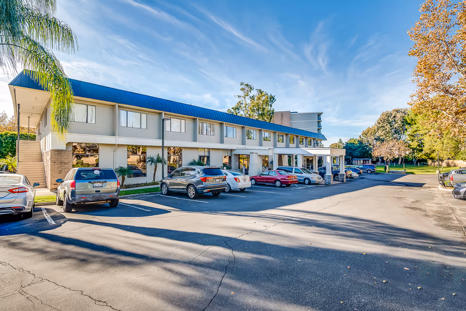 Exterior view of a two-story senior living facility building with a blue roof, several parked cars in front, palm trees, and a clear blue sky with some clouds.