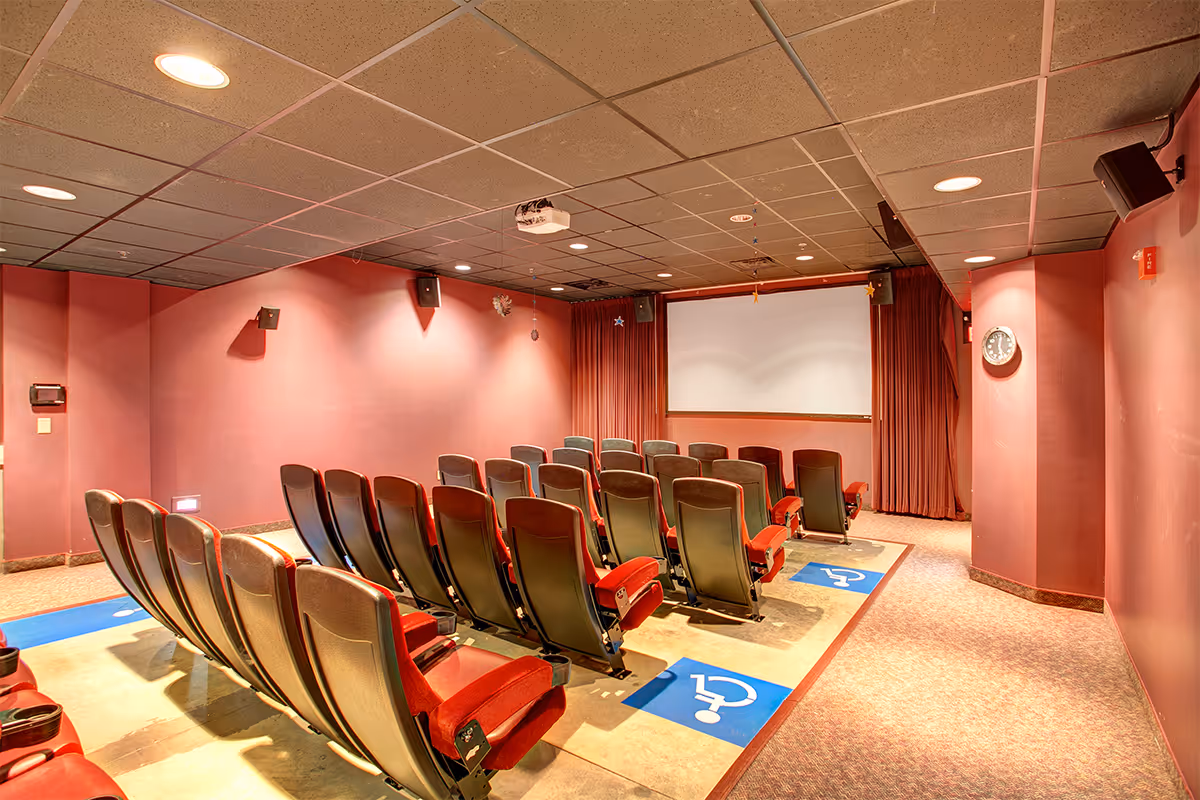 Interior view of a small theater room with rows of red cushioned seats facing a large white projection screen. The walls are painted a muted red color, and the ceiling has recessed lighting. There are blue handicap symbols on the floor indicating accessible seating areas.