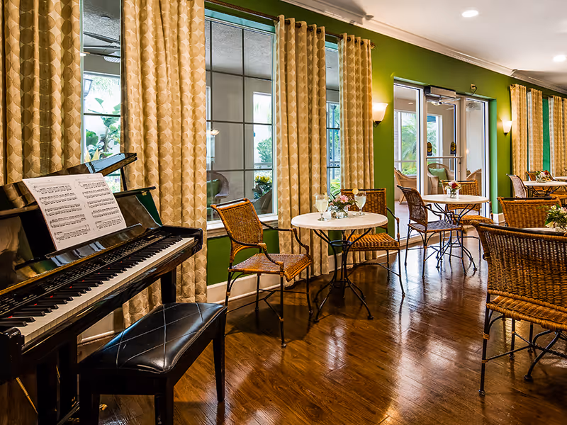 Interior view of a common area with green walls and large windows covered by beige patterned curtains. There are several round tables with wicker chairs arranged around them, each table decorated with small flower arrangements and glasses of water. A black piano with sheet music on the stand and a black cushioned bench is positioned on the left side of the room. The floor is polished wood, and there are wall-mounted lights providing warm illumination.