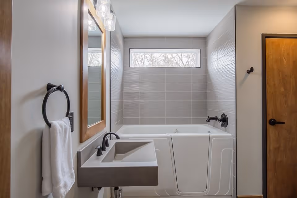 A modern bathroom featuring a walk-in bathtub with a door, a rectangular sink with black fixtures, a wooden framed mirror above the sink, a towel hanging on a black ring holder, a small horizontal window above the bathtub, and a wooden door on the right side.