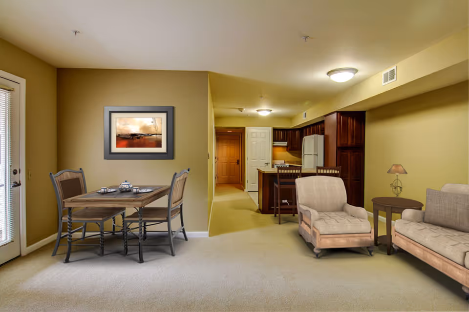 Interior view of a senior living facility apartment at Breeze Park showing a combined living and dining area with beige walls and carpet. The dining area has a wooden table set with four chairs and a tea set. The living area includes a cushioned armchair, a sofa, and a side table with a lamp. In the background, there is a kitchen with wooden cabinets, a refrigerator, and a small breakfast bar with two chairs. A framed abstract painting hangs on the wall above the dining table.
