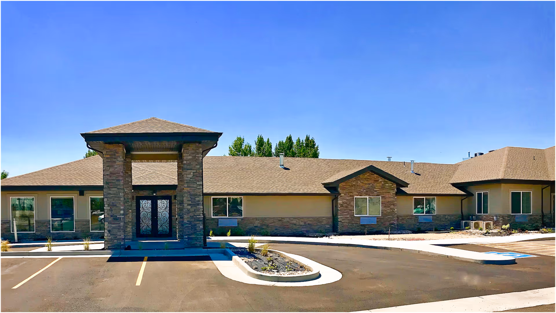 Front exterior view of Our House Assisted Living of Tremonton building with a covered entrance supported by stone pillars, multiple windows, and a parking area in front under a clear blue sky.