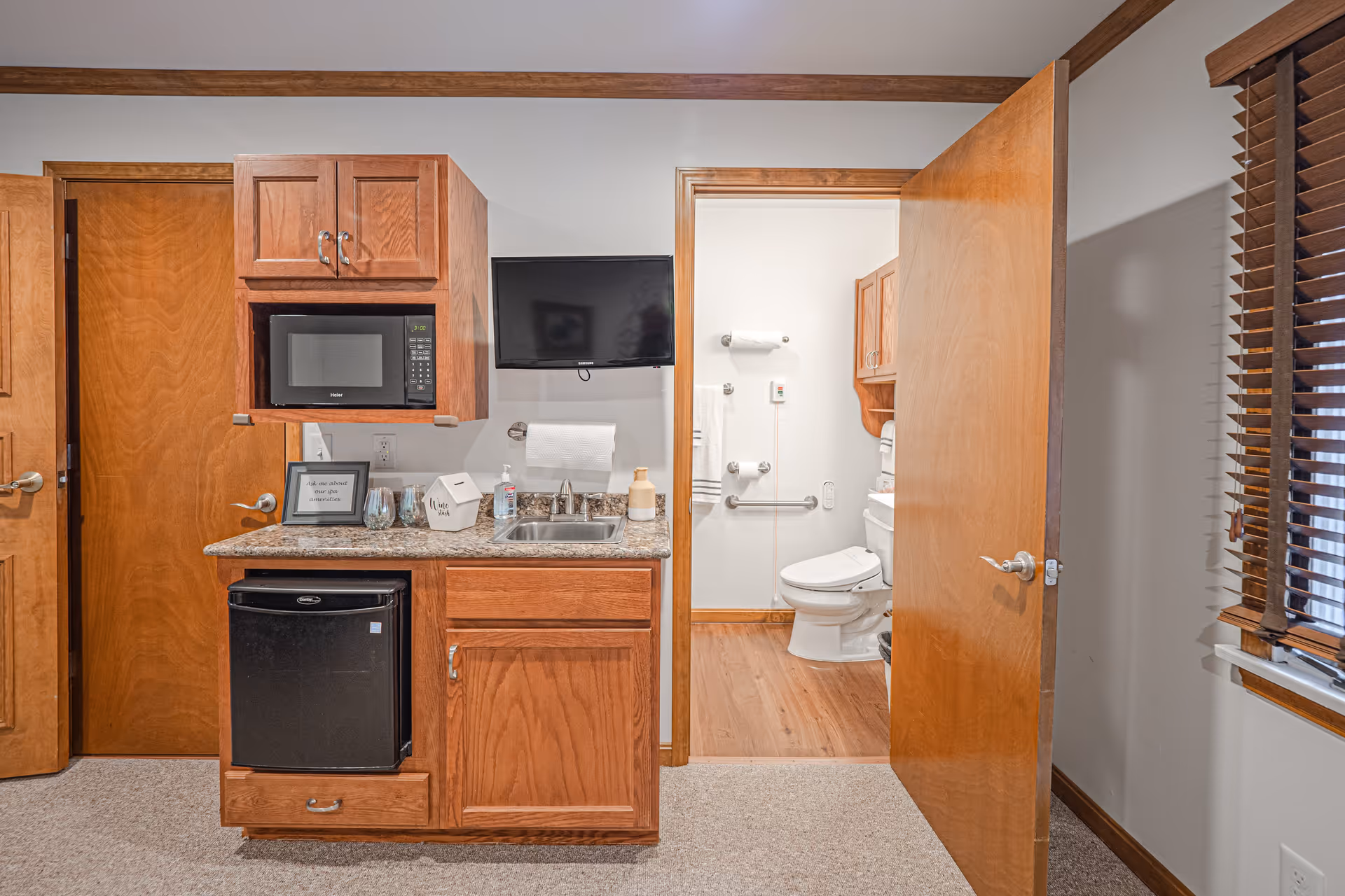 Interior view of a room in a senior living facility showing a kitchenette area with wooden cabinets, a microwave, a small refrigerator, a sink, and a mounted flat-screen TV. To the right, an open door reveals a bathroom with a toilet, grab bars, and wooden cabinetry. A window with wooden blinds is visible on the far right.