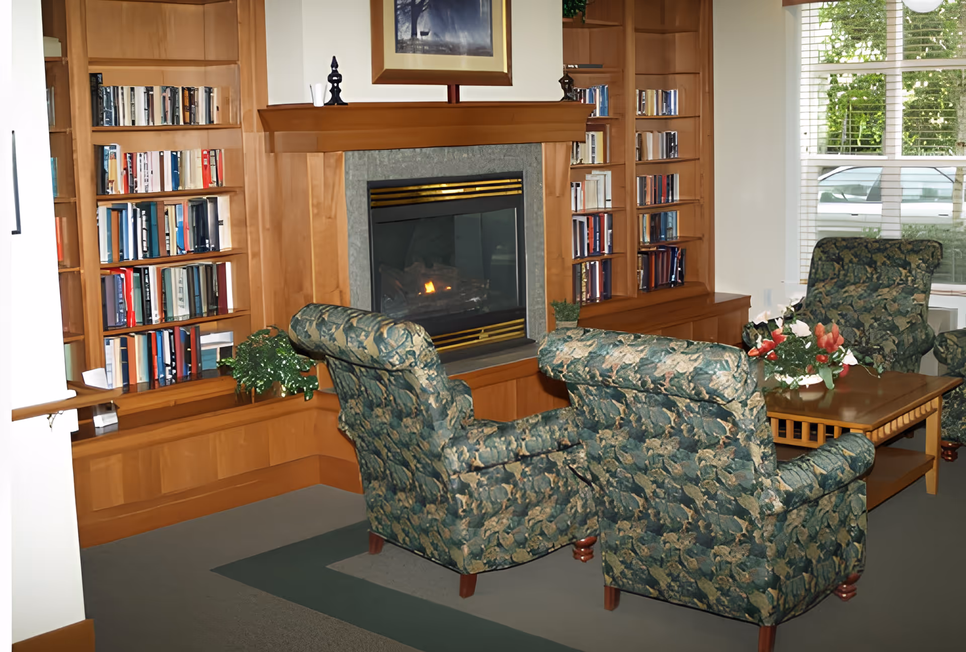 A cozy living room area with two floral-patterned armchairs facing a lit fireplace surrounded by wooden built-in bookshelves filled with books. A wooden coffee table with a floral arrangement is positioned between the chairs. A window with white blinds allows natural light to enter the room.