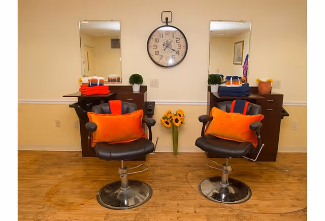 Two black salon chairs with orange cushions in front of two mirrors mounted on a beige wall. Each station has a small cabinet with folded towels and a small green plant. A round wall clock is centered between the mirrors above a vase with sunflowers on the floor.