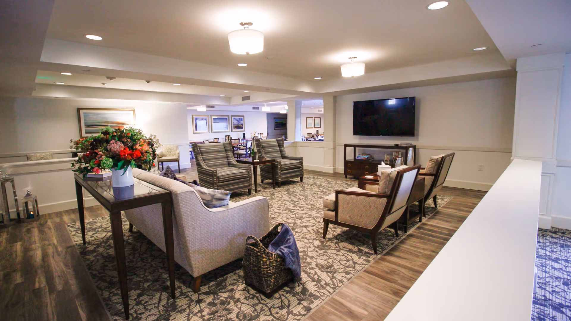 A spacious and well-lit living room area in a senior living facility with comfortable seating including a sofa, armchairs, and striped chairs arranged around a patterned rug. A flat-screen TV is mounted on the wall above a wooden console table. There is a table with a large floral arrangement and a basket with a blanket on the floor. In the background, a dining area with tables and chairs is visible, along with framed artwork on the walls.