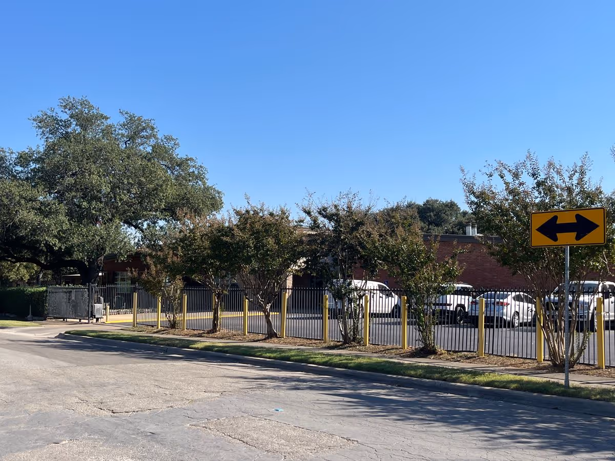 View of the facility's front exterior showing a fenced parking lot with parked cars, trees, and a two-way arrow sign under a clear blue sky.