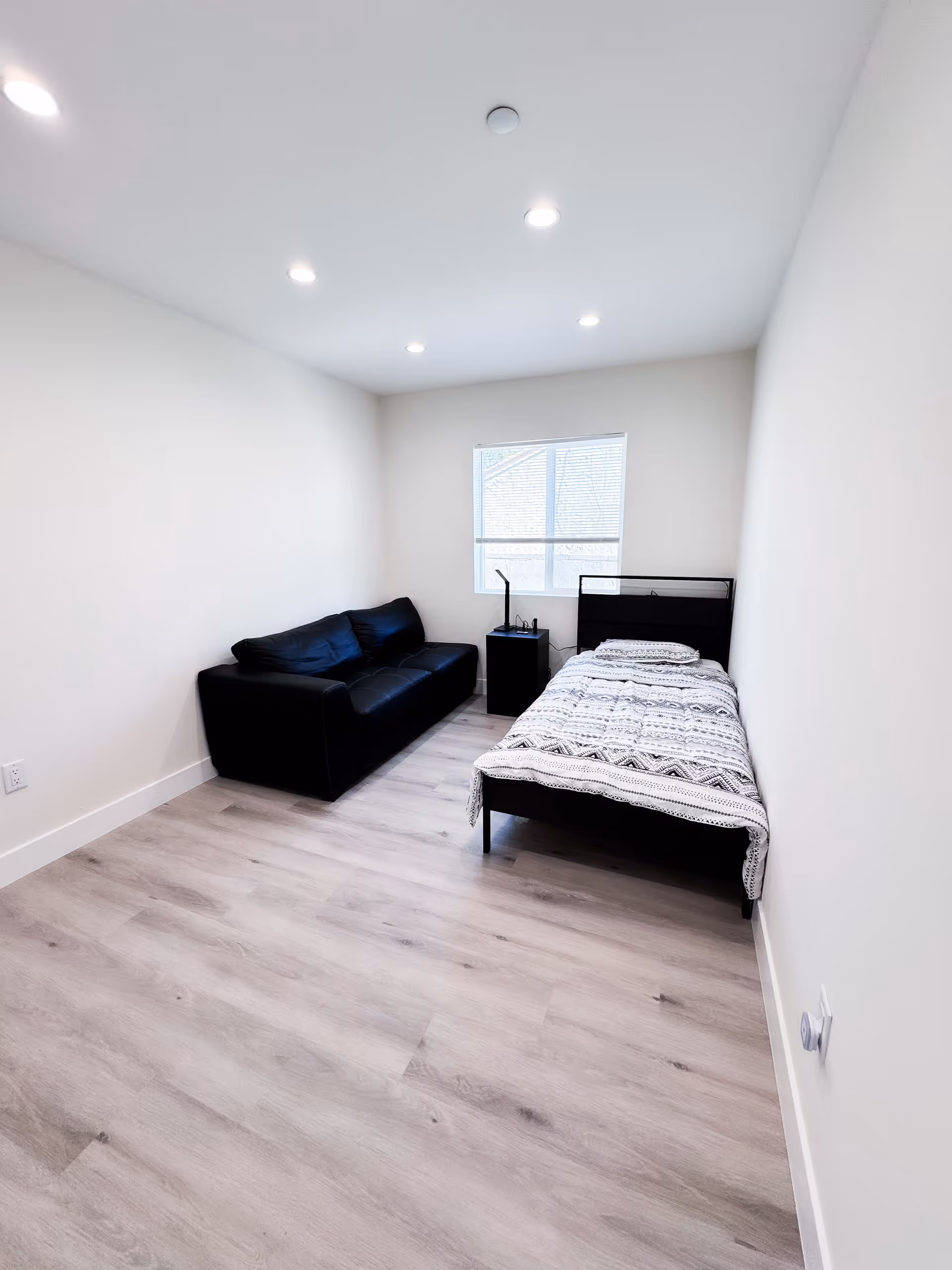 A simple bedroom with light-colored wooden flooring, white walls, and a window with blinds. The room contains a black leather couch, a single bed with a patterned blanket and pillow, and a small black nightstand with a lamp and some items on it. The ceiling has recessed lighting.