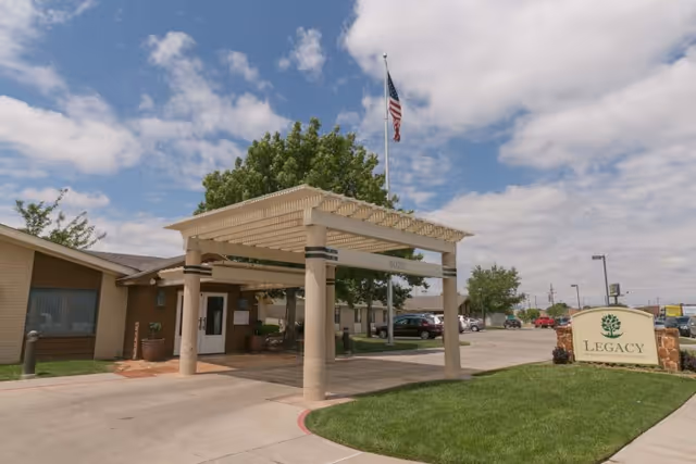 Exterior view of Legacy Rehabilitation And Living facility entrance with a covered drop-off area, an American flag on a flagpole, a green lawn, and a sign displaying the facility name.