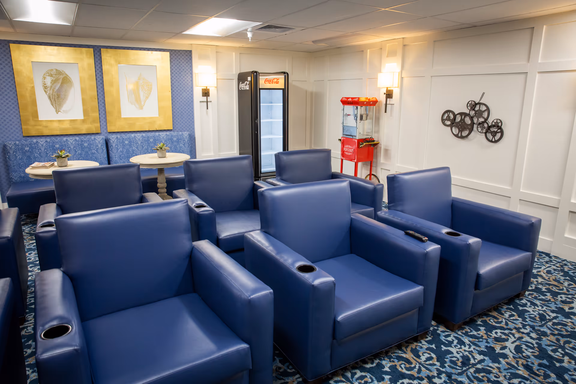 A cozy lounge area with six blue leather armchairs arranged in two rows on a patterned blue carpet. Behind the chairs, there are two small round tables with blue cushioned bench seating against the wall. The wall is decorated with two framed seashell pictures with gold borders, a popcorn machine, a Coca-Cola refrigerator, and a decorative wall piece resembling film reels. The room is softly lit with wall sconces.