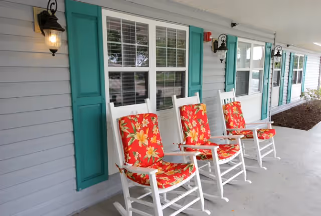 Covered front porch with four white rocking chairs with red floral cushions in front of a light gray building with teal shutters and wall lanterns.