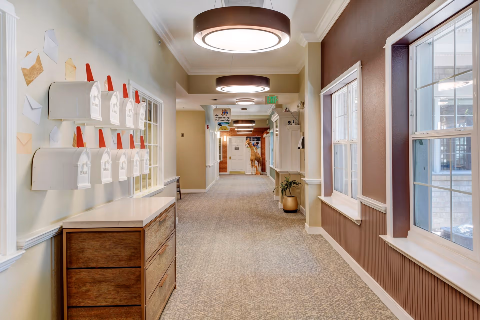 A well-lit hallway in a senior living facility with multiple white US mailboxes mounted on the left wall above a wooden cabinet. The hallway has beige walls, carpeted flooring, and large windows on the right side. Circular ceiling lights illuminate the corridor, and a decorative giraffe statue is visible near the end of the hallway.