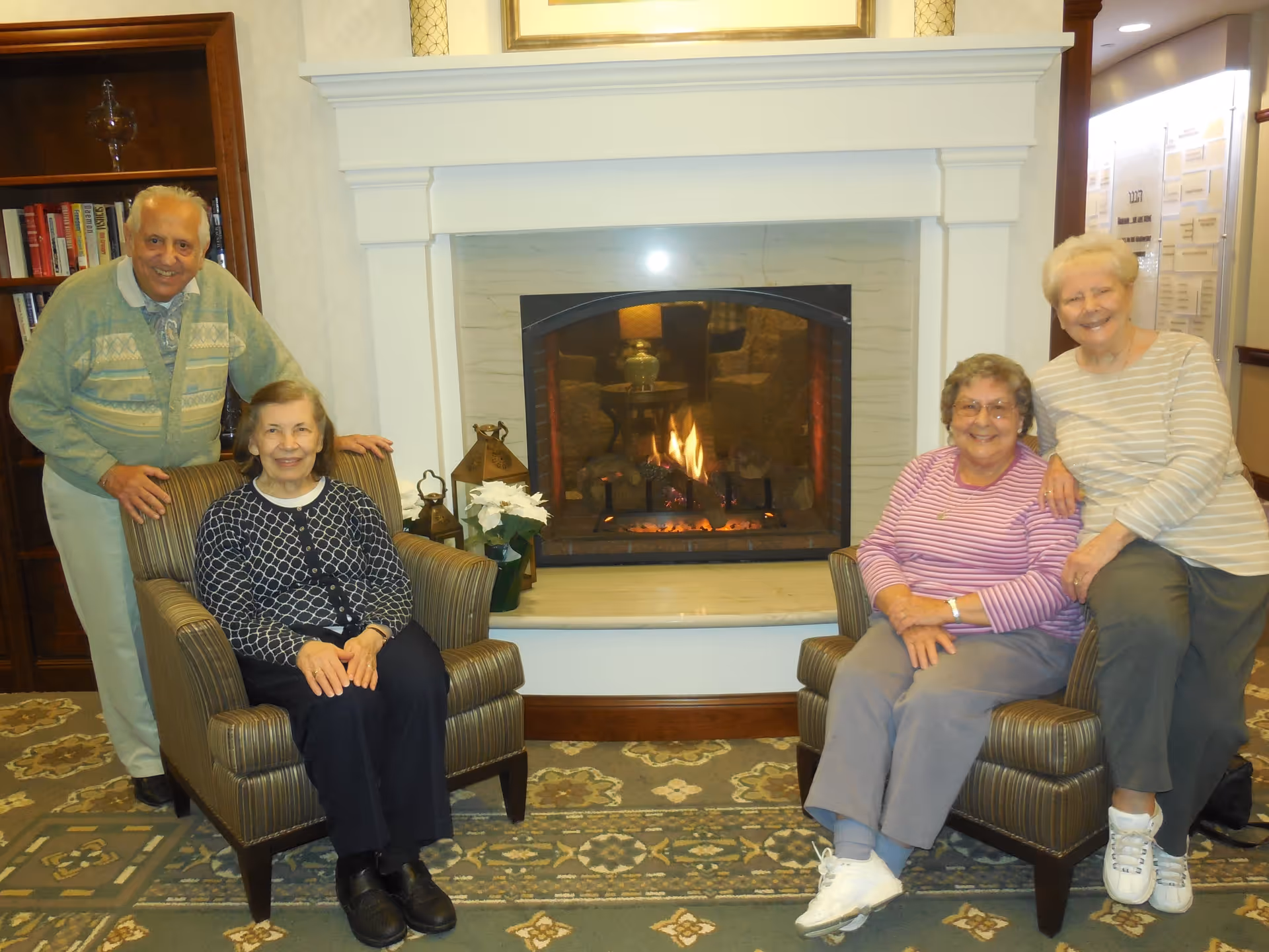 Four elderly individuals smiling and posing in a cozy living room area with a lit fireplace in the background. Two women are seated in armchairs, while a man and another woman stand behind them. The room features a patterned carpet, a bookshelf, and decorative lanterns near the fireplace.
