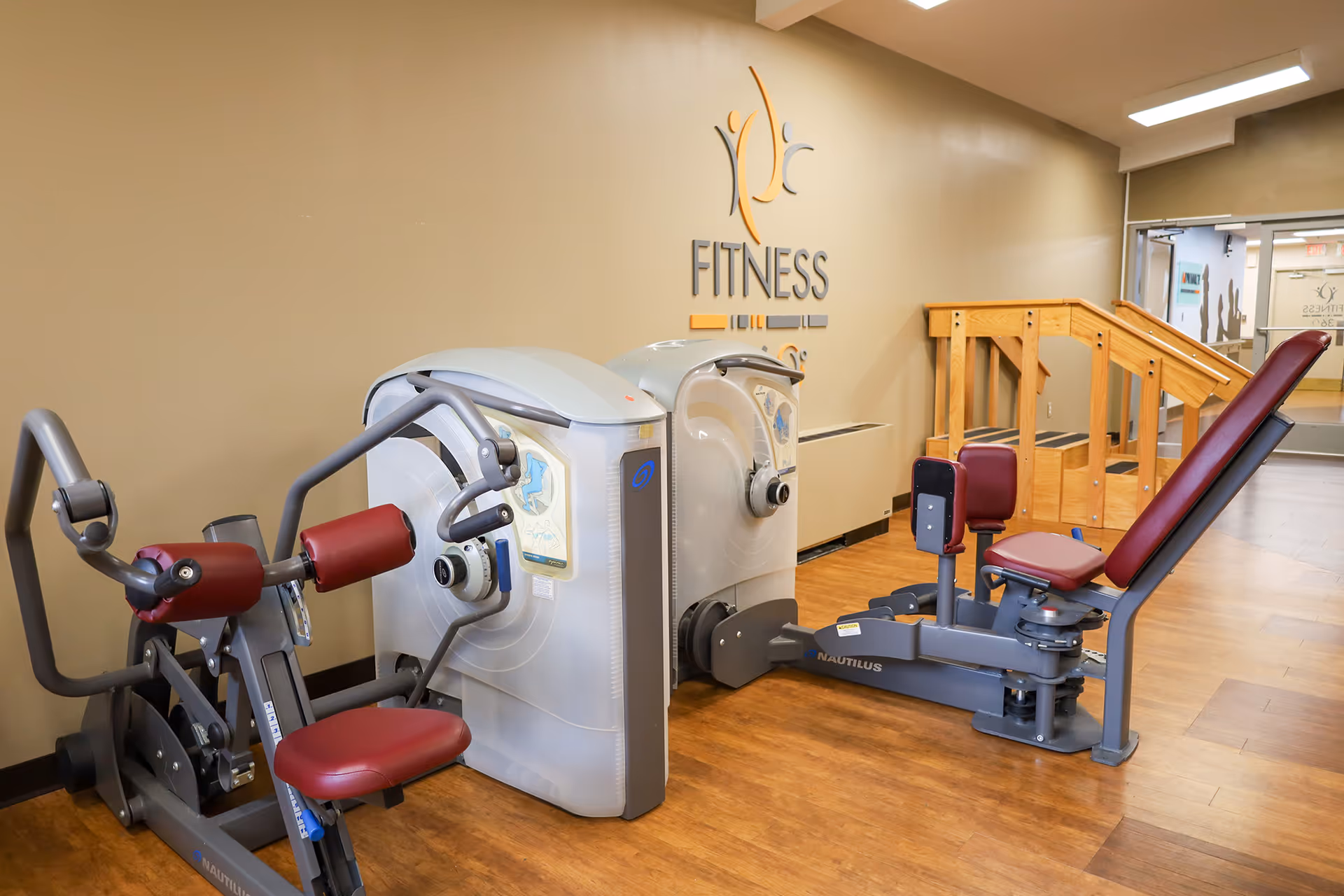 A fitness room with exercise machines including a seated leg press and an arm exercise machine, featuring maroon padded seats and handles. The room has wooden flooring and beige walls with a fitness center logo on the wall. There is a wooden ramp and a glass door in the background.