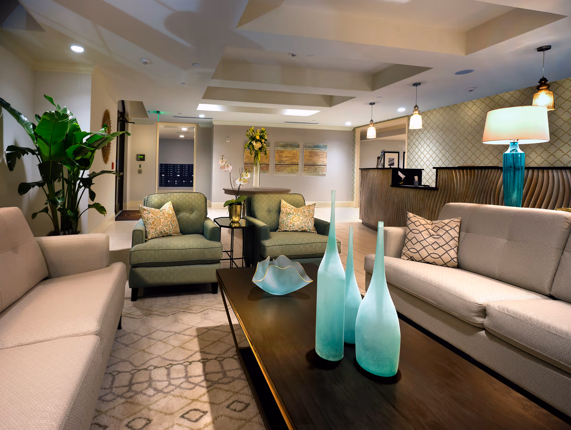 A well-lit, modern living room area in a senior living facility featuring two beige sofas and two green armchairs with patterned pillows arranged around a dark wooden coffee table. The coffee table holds three tall blue-green decorative vases and a matching bowl. The room has a patterned rug, a large green plant in the corner, a reception desk with pendant lights, and wall art in the background.