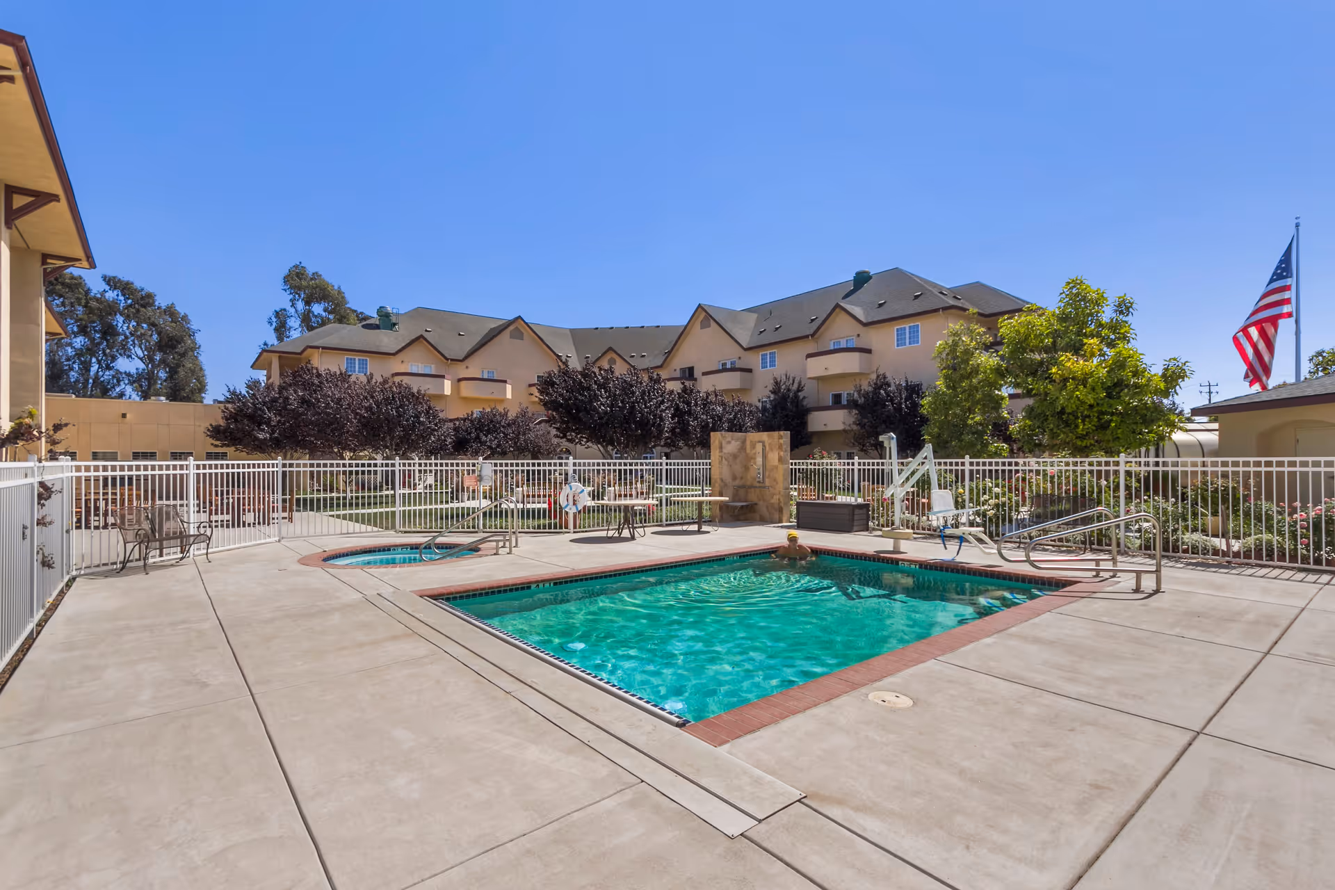 Outdoor swimming pool and hot tub area surrounded by a white safety fence with a multi-story residential building and trees in the background under a clear blue sky. An American flag is visible on the right side.