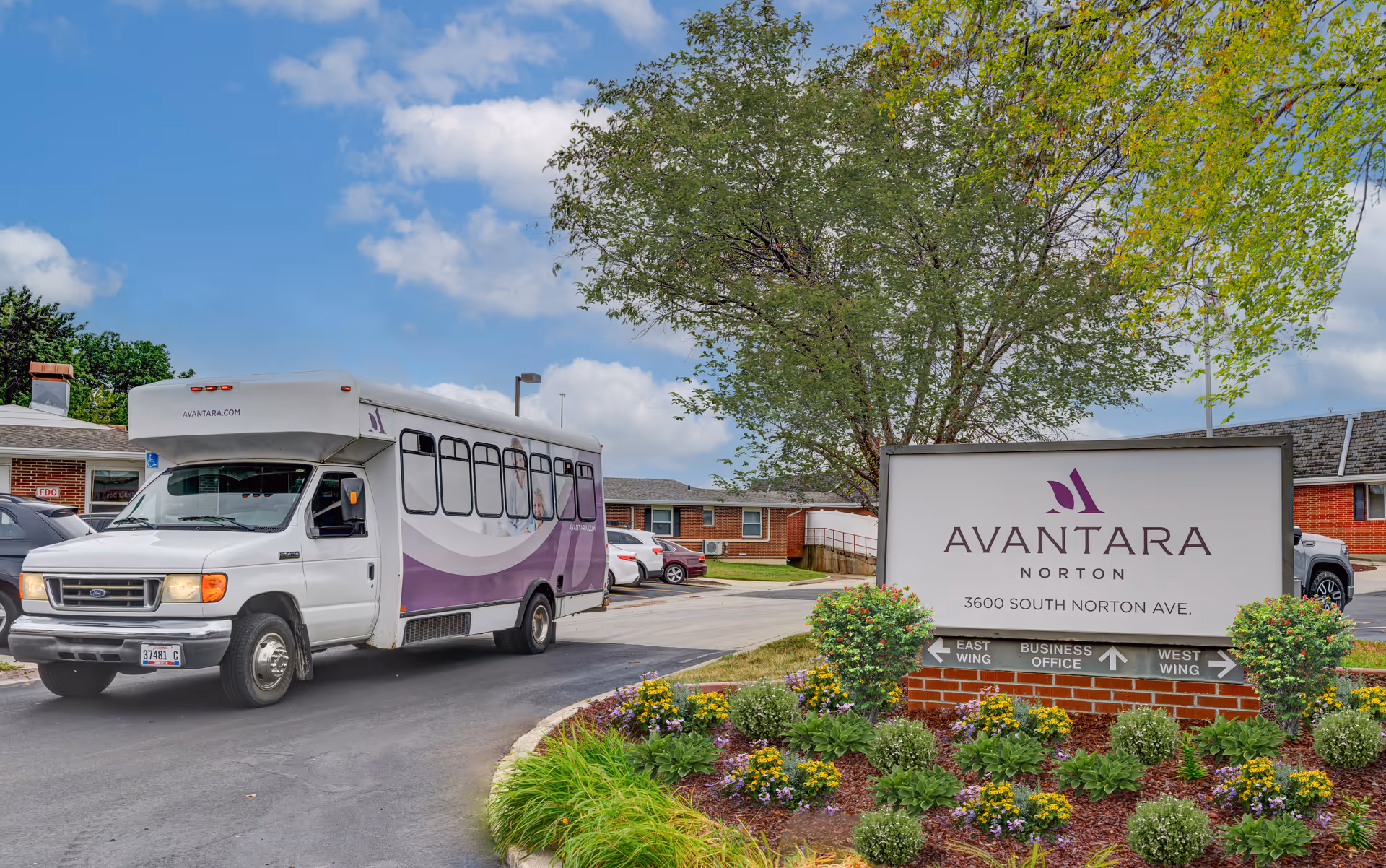 Exterior view of Avantara Norton senior living facility showing a white shuttle bus with purple accents driving on a paved road. To the right, there is a large sign with the facility name, address, and directional arrows for East Wing, Business Office, and West Wing. The area is landscaped with green shrubs and colorful flowers under a partly cloudy sky.