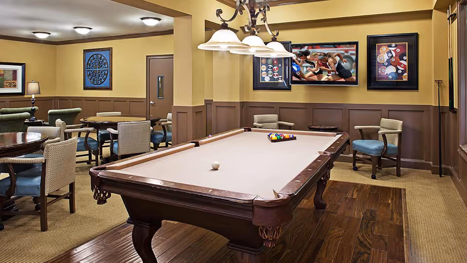 A game room with a pool table in the center, surrounded by several round tables and chairs. The walls are painted yellow with brown wainscoting, and there are framed pictures and a television mounted on the wall. The floor is a combination of carpet and wood, and there is a ceiling light fixture above the pool table.