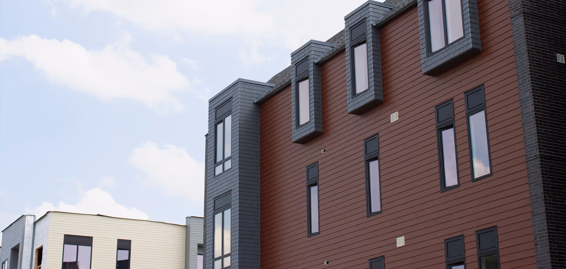 Exterior view of a modern multi-story building with reddish-brown siding and vertical windows under a partly cloudy sky.