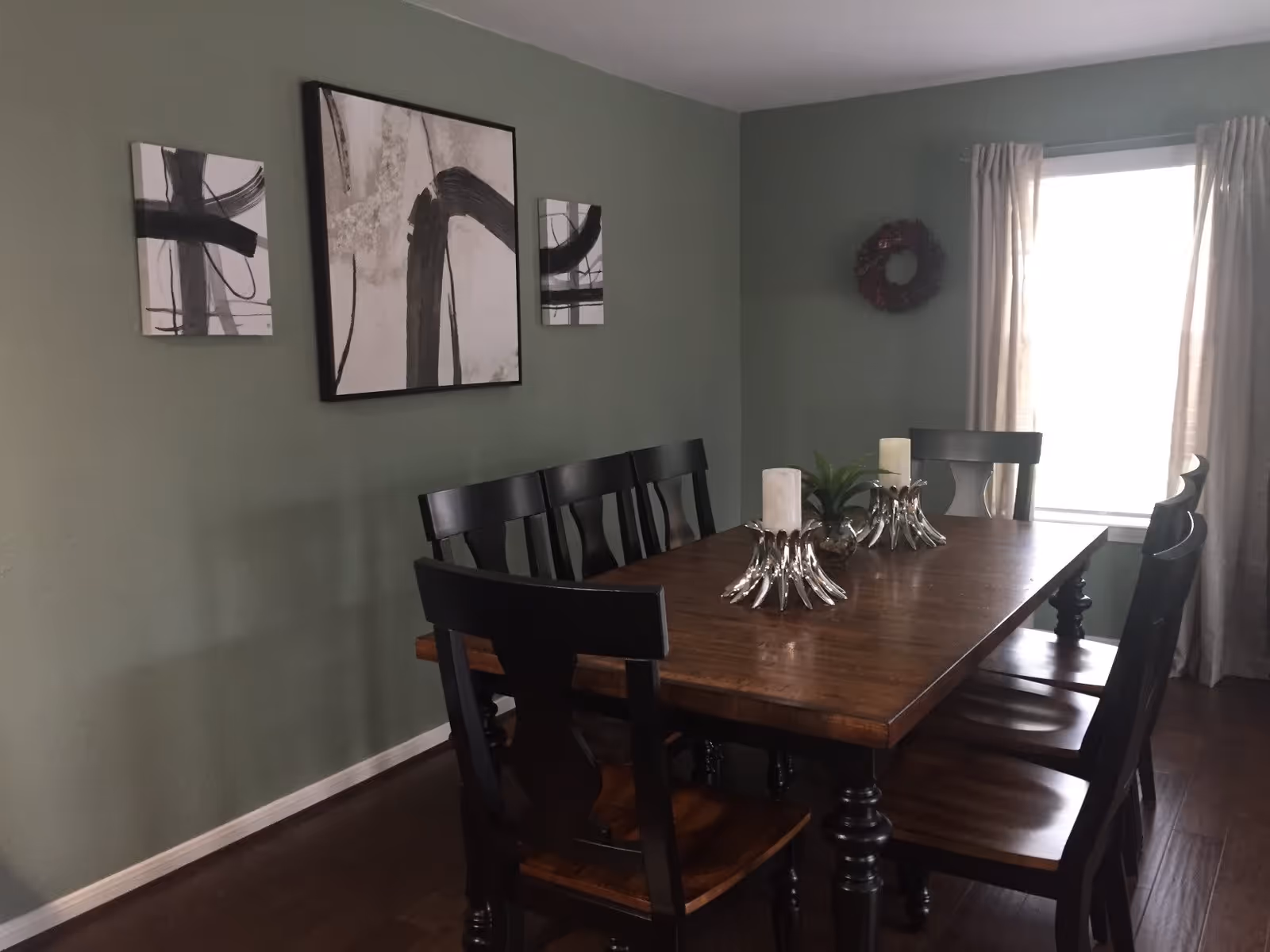 A dining room with a wooden table and eight black chairs. The table has decorative items including two candle holders and a small plant. The walls are painted green and decorated with three abstract black and white paintings. A window with light curtains is on the right side, letting in natural light.
