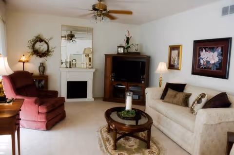 A cozy living room with a beige sofa adorned with decorative pillows, a red armchair, a wooden coffee table with a candle centerpiece on a small rug, a wooden entertainment center with a TV, a white fireplace with a mirror above it, and various lamps and framed artwork on the walls.