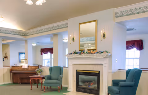 A cozy living room area featuring a white fireplace with a gold-framed mirror above it, flanked by two wall sconces. There are two teal upholstered armchairs positioned near the fireplace, a small wooden side table, and a piano against the wall with a floral arrangement on top. The room has large windows with red valances and a green carpeted floor.