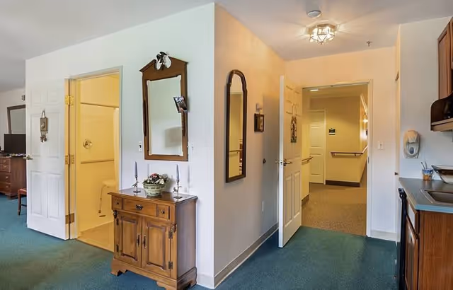 Interior view of a senior living facility room showing a small wooden cabinet with a mirror above it, a hallway with open doors leading to other rooms, a kitchen counter with a sink on the right, and a bathroom visible through an open door on the left.