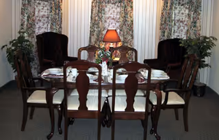 A dining room with a wooden dining table set for six people, including plates, cups, and a floral centerpiece. The room has three tall windows with floral curtains, two dark upholstered armchairs, and two potted plants on either side of the windows. A table lamp with a red shade is placed on a small table behind the dining table.