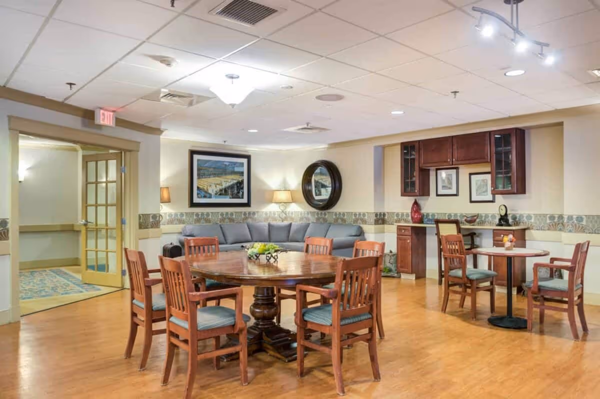 A cozy common area in a senior living facility featuring a round wooden table with six chairs, a smaller round table with three chairs, a gray sectional sofa against the wall, framed artwork, a round mirror, and built-in wooden cabinetry with decorative items. The room has wood flooring, soft lighting, and an exit door with glass panels.