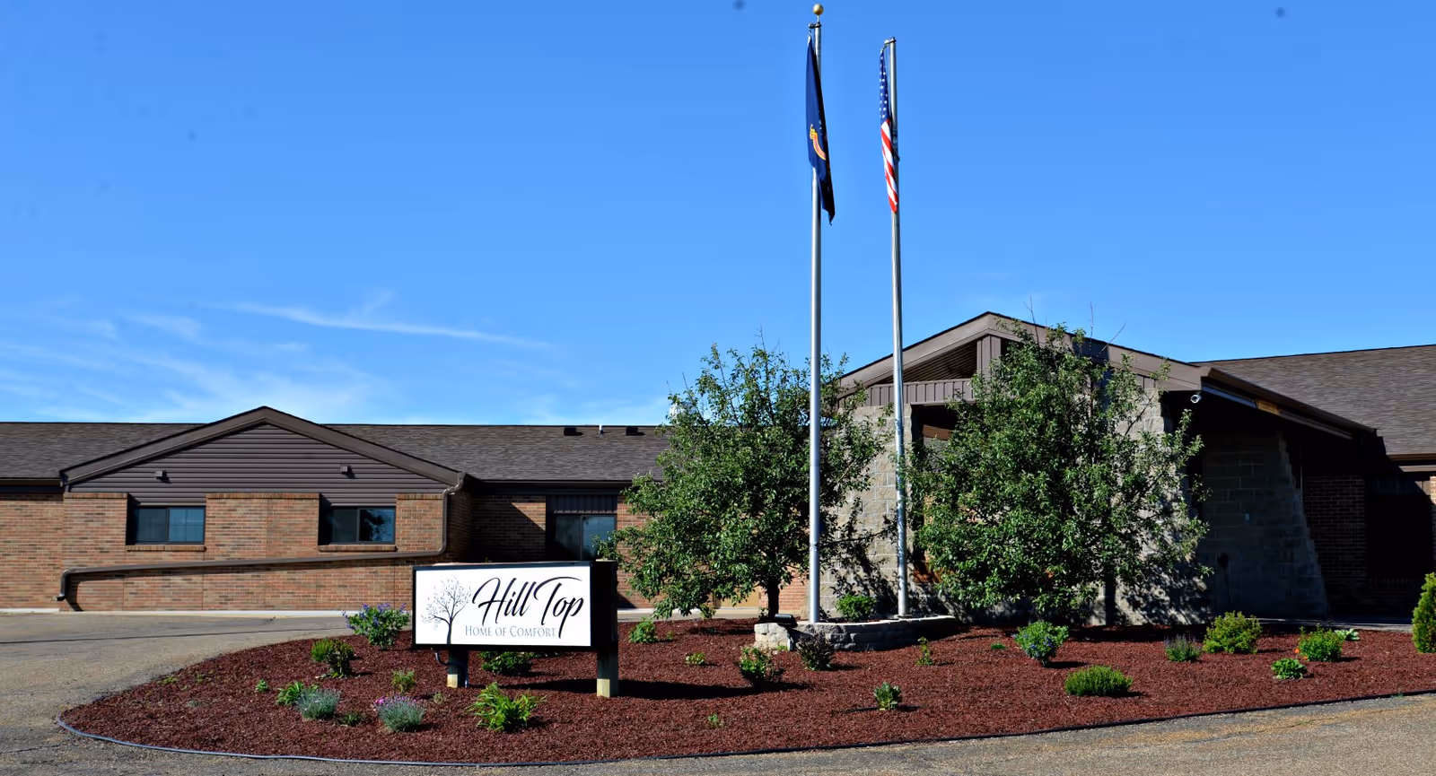 Exterior view of Hill Top Home Of Comfort building with a landscaped garden area in front, two flagpoles with flags, and a clear blue sky.