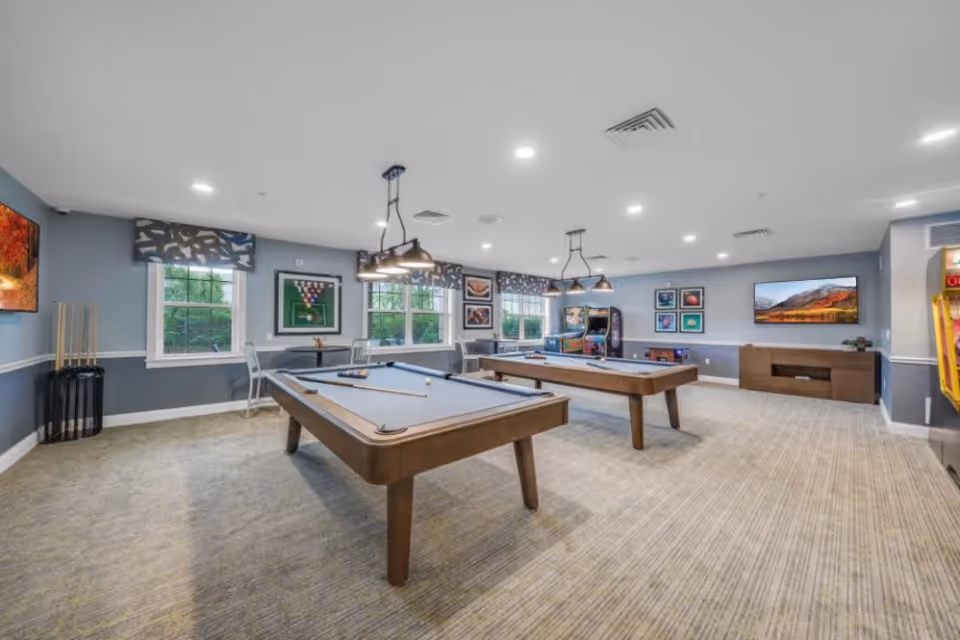 A spacious game room with two pool tables, several chairs, and arcade machines along the far wall. The room has large windows with patterned valances, framed artwork on the walls, and ceiling lights. The floor is carpeted and the walls are painted gray with white trim.