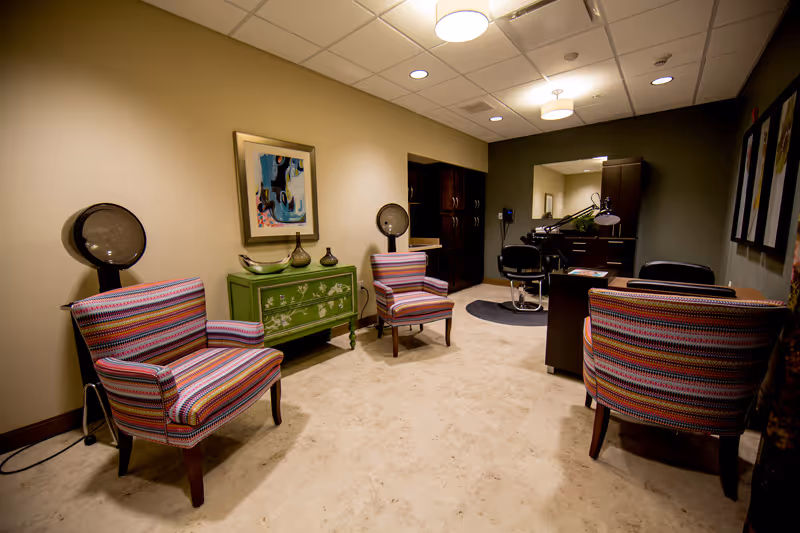 Interior view of a salon or beauty room with three striped armchairs, a green decorative cabinet with vases, two hair dryer chairs, a large mirror, and dark wood cabinetry under warm ceiling lights.
