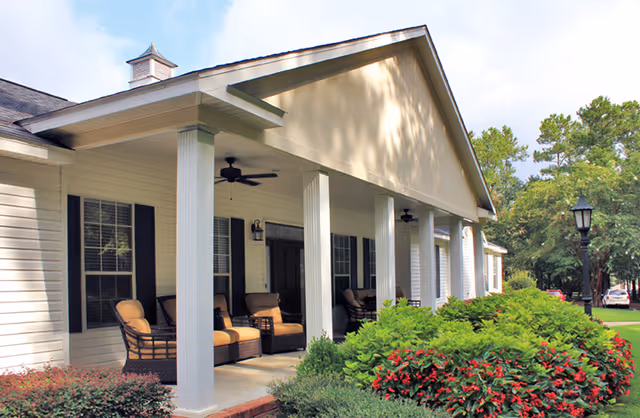 Covered front porch of a senior living facility with white columns, wicker chairs, ceiling fans, and landscaped shrubs.