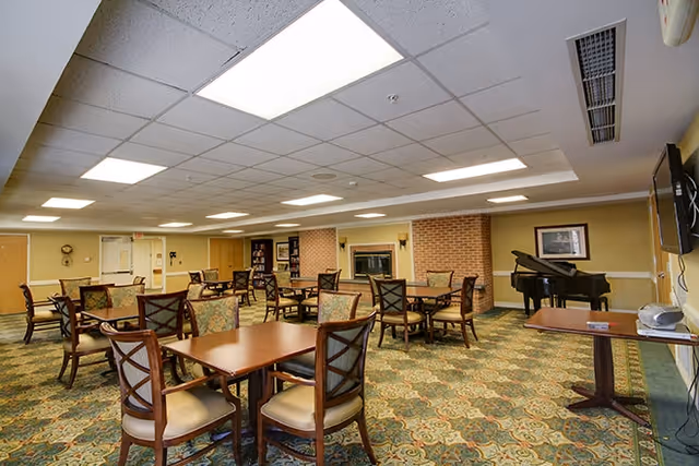 A spacious common area with multiple wooden tables and cushioned chairs arranged on a patterned carpet. The room features a brick fireplace, a grand piano, a wall-mounted television, and soft overhead lighting in a drop ceiling.