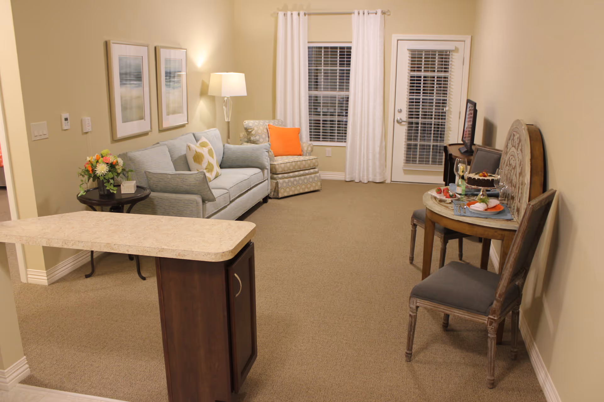 A cozy living room area with a light blue sofa adorned with patterned pillows, an armchair with an orange pillow, a floor lamp, and two framed pictures on the wall. There is a small round table with a flower arrangement next to the sofa. On the right side, a small round dining table is set with a cake, wine glasses, and plates, accompanied by two chairs. The room has beige walls, carpeted floor, a window with white curtains, and a door with blinds.
