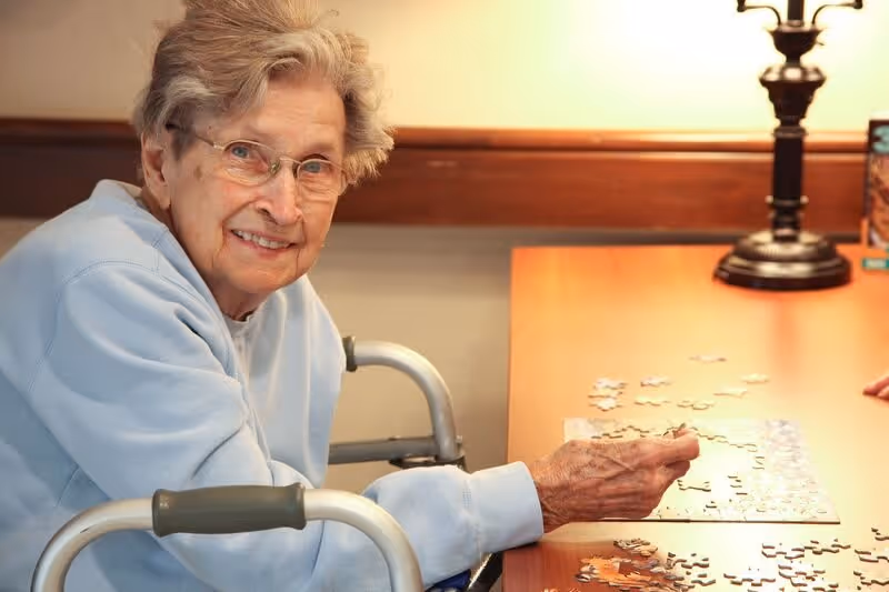 An elderly woman with glasses and gray hair is sitting at a wooden table working on a jigsaw puzzle. She is smiling and wearing a light blue sweater. A lamp is visible on the table in the background.