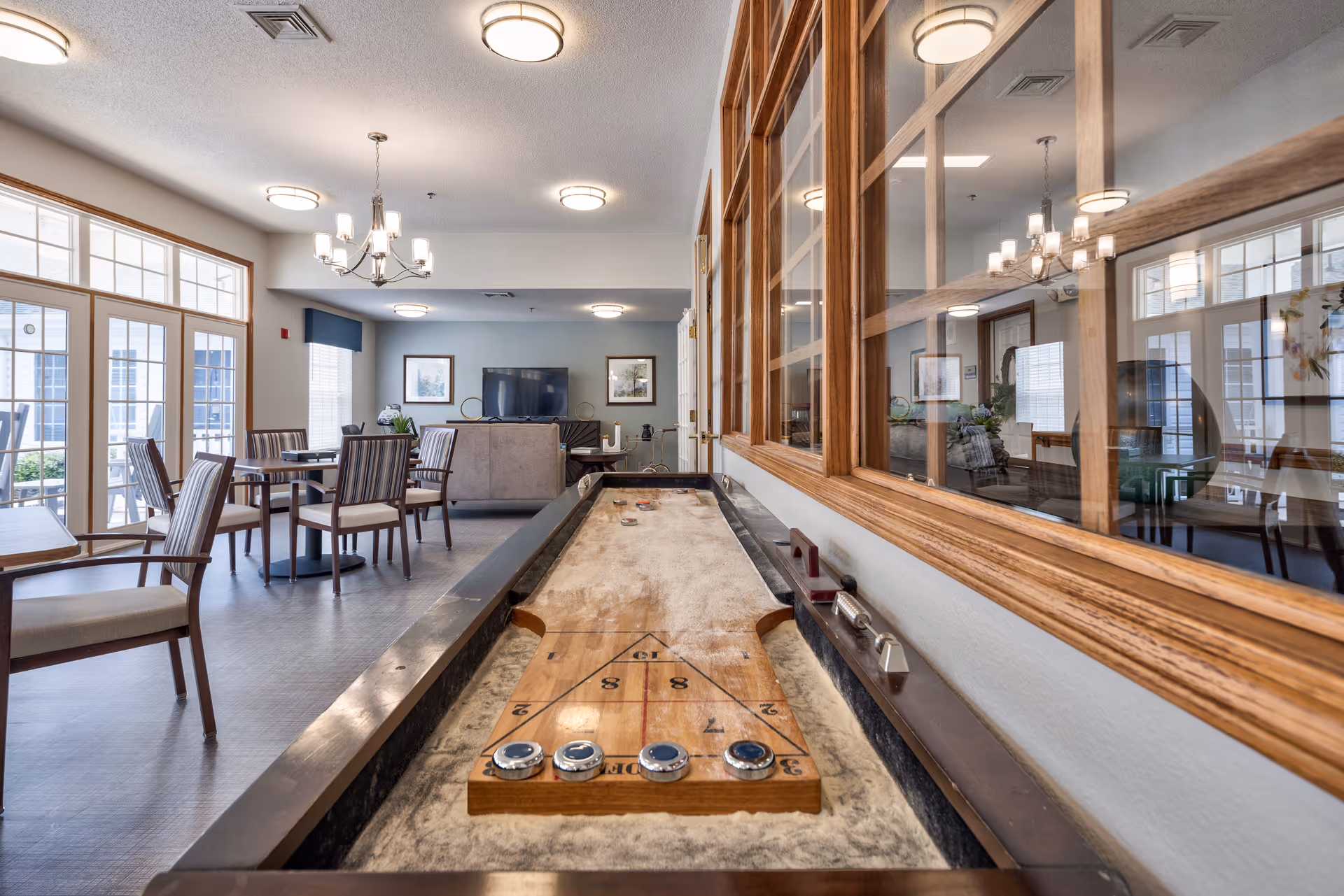 Interior view of a senior living facility common area featuring a shuffleboard table in the foreground, several tables and chairs for seating, large windows allowing natural light, a television mounted on the far wall, and decorative framed pictures. The room has a bright and welcoming atmosphere with ceiling lights and a chandelier.