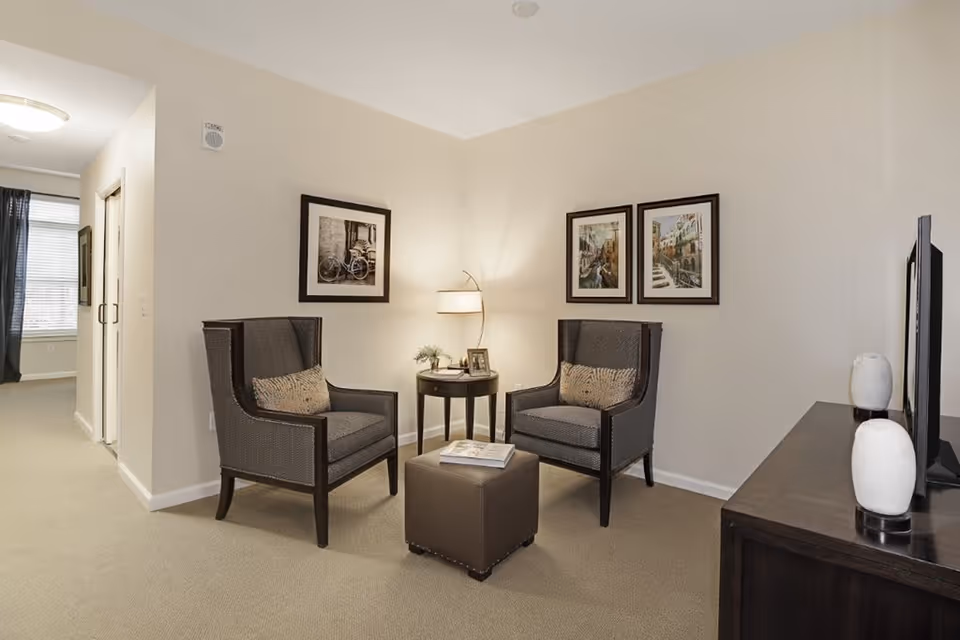 A cozy living room area with two dark upholstered armchairs facing each other, a small round side table with a lamp and decorative items between them, and a dark wooden TV stand with two white vases. The walls are light-colored and decorated with three framed pictures. A hallway with a window and dark curtains is visible in the background.