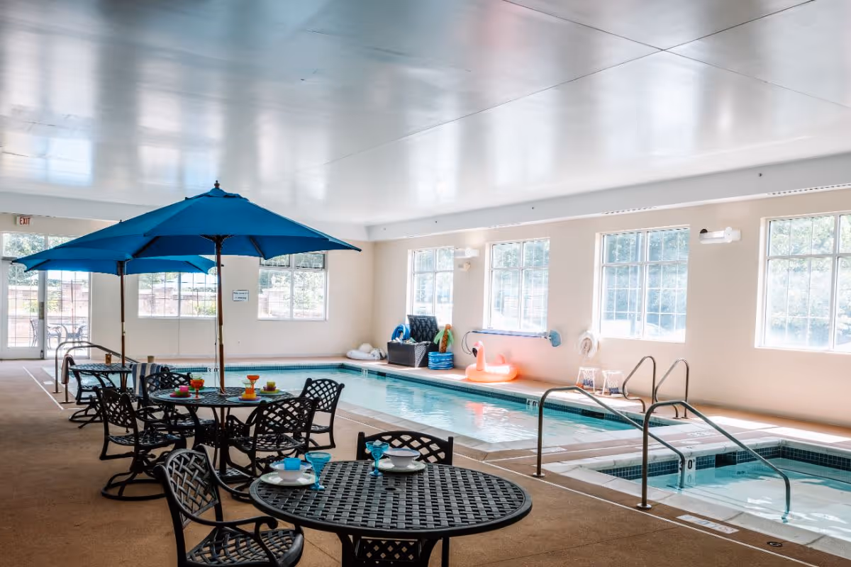 Indoor swimming pool area with several black metal tables and chairs, each table shaded by a blue umbrella. The pool is rectangular with a smaller hot tub adjacent to it. Large windows line the walls, allowing natural light to fill the space. Pool toys and flotation devices are visible near the far wall.