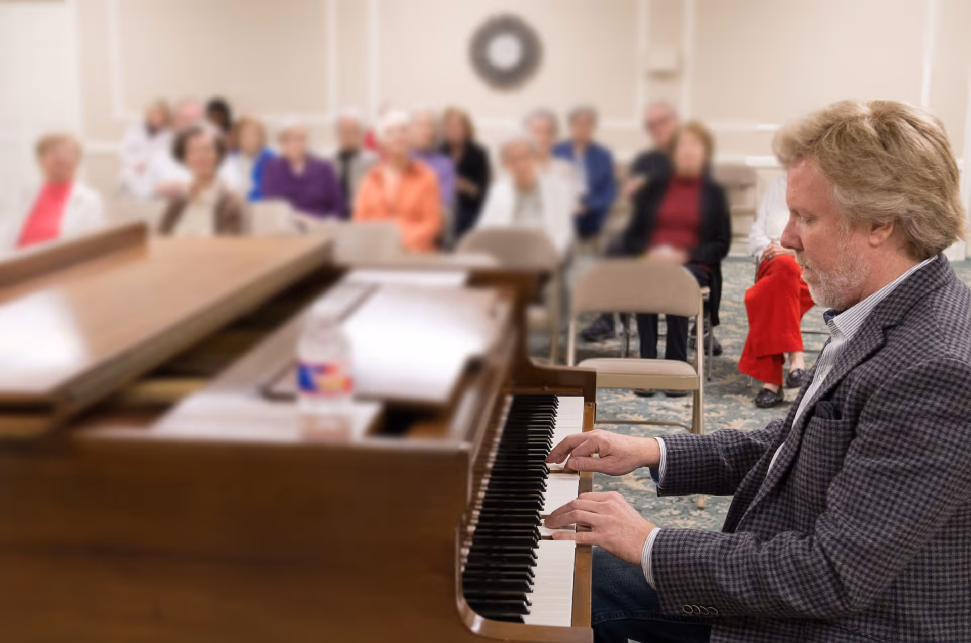 A man with light hair and a beard is playing a piano in a room with an audience of elderly people seated on chairs, attentively watching the performance.